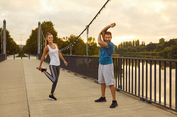 Two happy people having a fitness workout in the city. Young woman and man in sportswear standing on the bridge and doing warming up stretching exercises for arm and leg muscles. Sport concept
