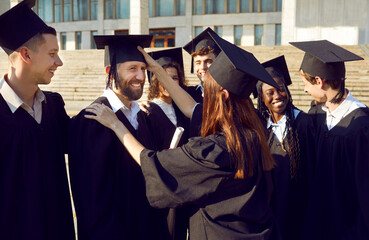 Student girl puts black academic hat on head of happy young man. Group of joyful college or university friends wearing graduate caps and gowns celebrating their graduation day and having fun together - obrazy, fototapety, plakaty