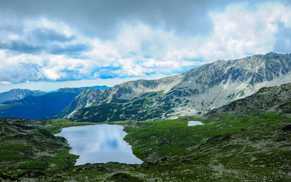 Rainy clouds above the mountain peaks. A glacial lake is in a grassy ravine, and laying in the shadows. Sunlight lits the nearby mountain pastures. Bucura Lake, Carpathia, Romania.