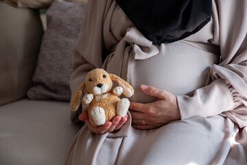 Pregnant female tummy in 8th month holding Stuffed rabbit toy while sitting on couch