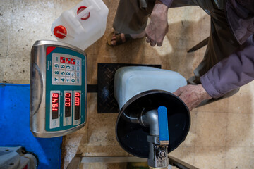 An old man is filling fresh olive oil into plastic gallons using a funnel and weighing scale in a...