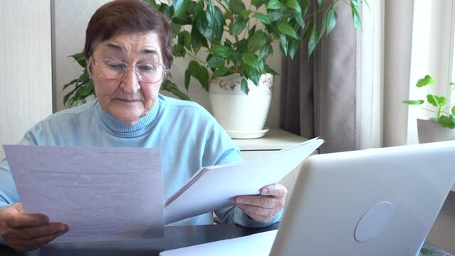 An elderly woman organizes her home office, carefully reviewing papers, bills, and pension insurance documents. She compiles a will. Ideal for content about active and organized lifestyle of seniors.