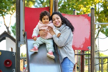 Dominican mother playing with her toddler daughter at the slide in a park. Latin family.