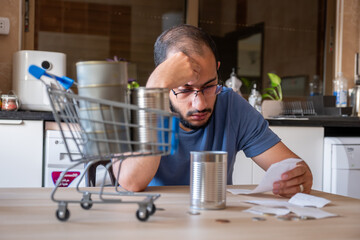 man sitting in the kitchen holding receipt feeling frustrated due inflation and increase of daily...
