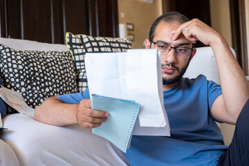 Young Male sitting in the living room checking receipts and using calculator to find the amount of...