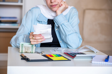 Young Female wearing hijab sitting in the living room checking receipts and using calculator to...