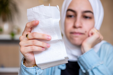 Young Female wearing hijab sitting in the living room checking receipts and using calculator to...