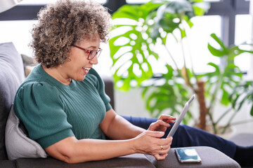 Beautiful middle-aged woman using a computer tablet to read a book and surf internet. Modern technologies used by elderly people. Mature woman using digital tablet at home - obrazy, fototapety, plakaty