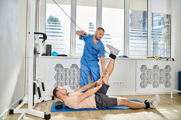 experienced recovery expert assisting man working out on exercise machine in gym of kinesio center - obrazy, fototapety, plakaty