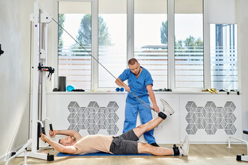 skilled rehabilitologist assisting man working out on exercise machine in gym of kinesio center