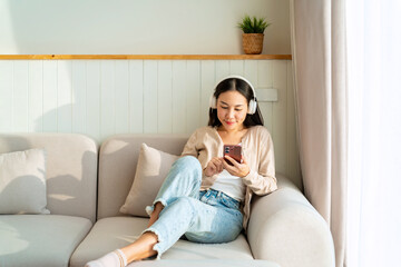 Obraz premium Happy young Asian woman with mobile phone listening music in headphones and relaxing on safa at home