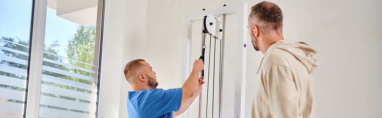 rehabilitologist in blue uniform showing training equipment to man in kinesio center, banner