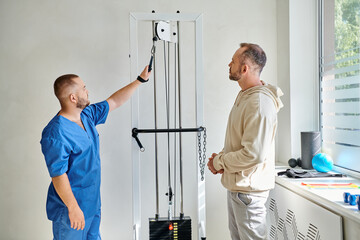 professional rehabilitologist in blue uniform showing training machine to man in kinesiology center - obrazy, fototapety, plakaty