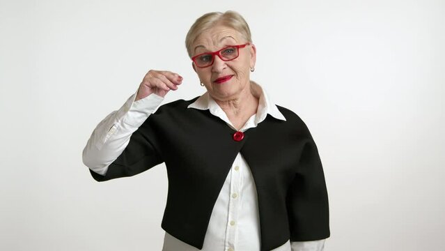 Old woman with short blonde hair, red lipstick, wearing white shirt, black jacket with big red button standing over white background, shrugging shoulders. High quality 4k footage