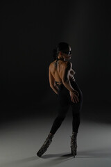 Young woman in black open clothes poses on black background of backlit studio. The dancer demonstrates elements of dance choreography in high heels. The concept of the dance promo shot.