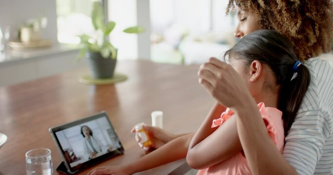 Biracial mother with sick daughter at home, having tablet video call with female doctor, slow motion