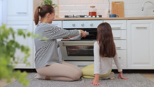 Smiling mother and daughter taking baking out of oven sitting near oven smelling delicious sweets kid helping mother on kitchen family posing at home while sitting on the floor.