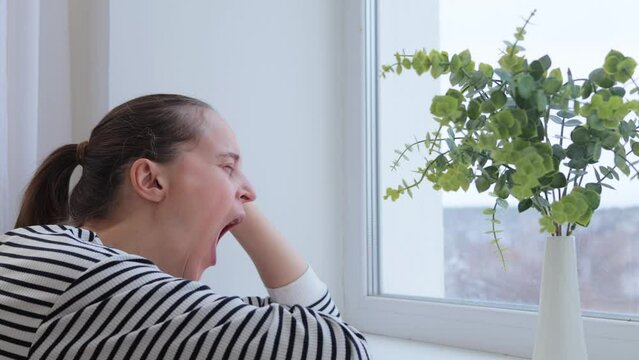 Adult Caucasian woman with ponytail with boring face looking out of window and yawning being sad bored and sleepy leaning on her head waiting for something