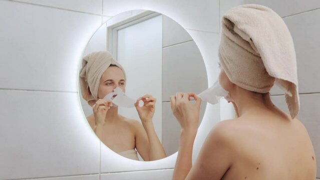Winsome beautiful Caucasian woman wrapped in towel on her head looking at mirror at her reflection removing cloth mask doing home cosmetology procedures standing in bathroom