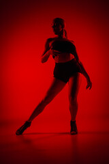 Fototapeta na wymiar Silhouette female dancer in black shorts and top. Young woman poses gracefully and shows off slender flexible body in dark studio with red light. Elements of high heels dance choreography.
