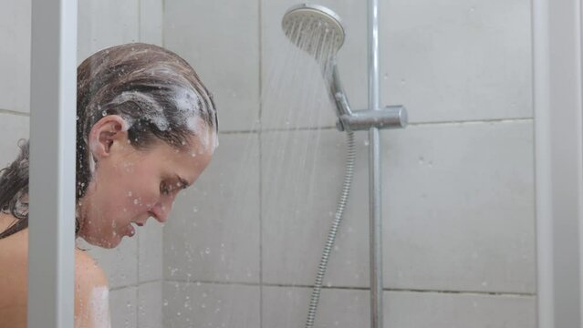 Funny Caucasian bare young adult brown haired woman reacting while taking cold shower waiting for hot water while standing in foam on her hair