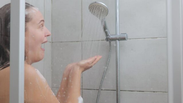 Shocked Caucasian bare young woman reacting while taking cold shower grimacing from freeze water standing with foam on her head cold surprise during morning hygiene routine