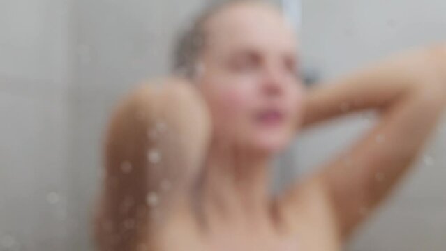 Home spa. Beauty routine. Relaxing shower. Daily skincare. Young adult woman washing her hair and body in cabin behind steam blurred glass with water drops