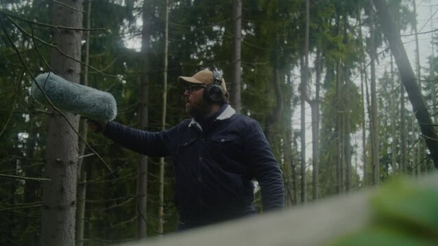 Sound technician wearing headphones records sounds of nature for movie in coniferous wood using professional audio equipment. Caucasian man works with furry windjammer microphone outdoors. Filmmaking.