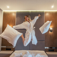 Woman having fun and jumping with bathrobe on bed in hotel room