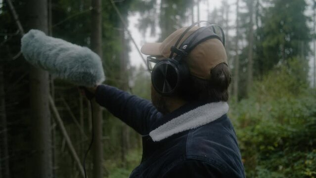Male tourist wearing headphones records sounds of nature for movie in coniferous wood using furry windscreen microphone. Sound engineer works with professional audio equipment outdoors. Film industry.