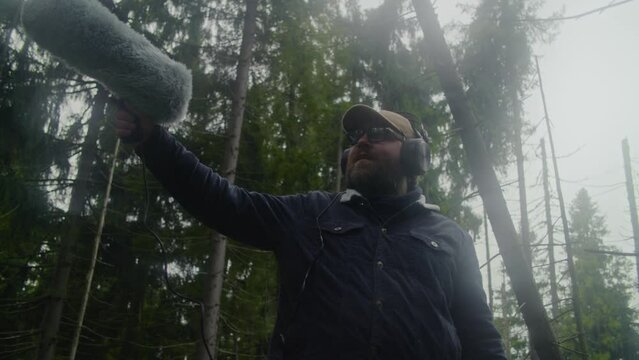 Caucasian man wearing headphones records sounds of nature for movie in coniferous wood using furry windscreen microphone. Sound engineer works with professional audio equipment outdoors. Low angle.