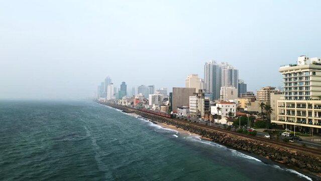 Aerial footage presents Colombo skyline oceanfront, urban landscape, Sri Lanka. High-rises flank coastline train, traffic move parallel to beach. Tourism, real estate promo potential.