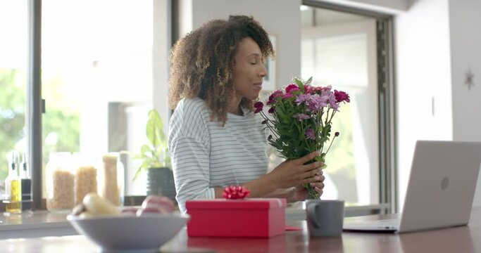 Happy biracial woman with gift and flowers having video call on laptop at home, slow motion