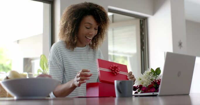 Happy biracial woman with gift and flowers having video call on laptop at home, slow motion