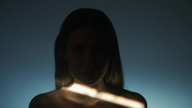 Portrait of female model in studio. Silhouette of appealing woman face looks at camera on low light background, ray of light moves up the face