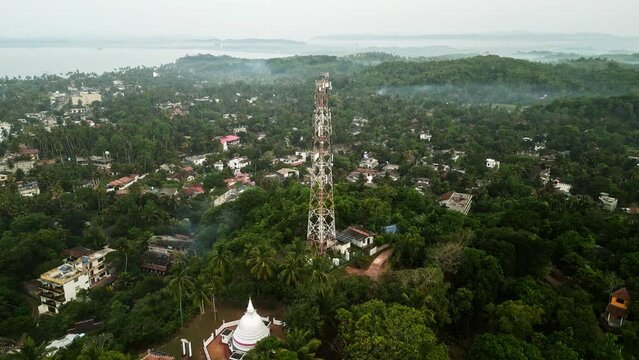 Aerial view of towering communications mast amidst lush greenery on island ensuring mobile network connectivity, broadcasting services to area with coastal backdrop, morning evening ambient light.