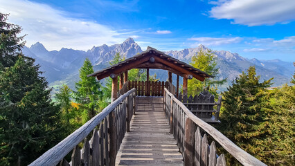 Fototapeta na wymiar Wooden viewing platform connected by bridge with vistas to majestic mountain ranges of untamed Sexten Dolomites, South Tyrol, Italy. Lookout from mount Helm (Monte Elmo), Carnic Alps, Austria border