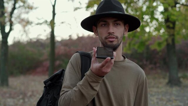 Technology and people concept. Handsome young man wear black hat walking in autumn park and using voice command recorder on smartphone