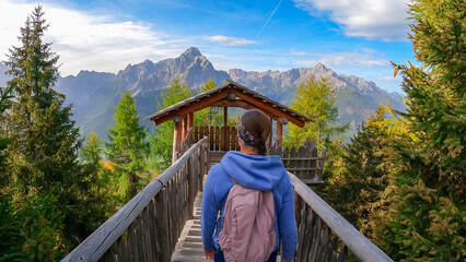Hiker woman with backpack on wooden viewing platform with panoramic view of majestic mountain peaks of untamed Sexten Dolomites in South Tyrol, Italy, Europe. Hiking concept in Italian Alps in fall - obrazy, fototapety, plakaty