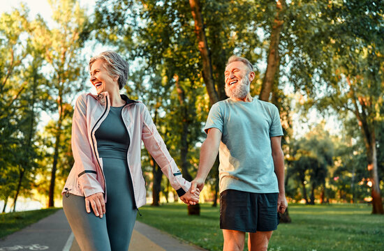 Modern active cheerful sports couple of senior people holding hands and talking cheerfully outdoors together. The woman turned her face to the rays of the sun and laughs. The joy of life.