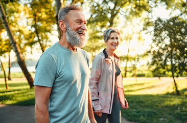 Active hobby on retirement. Grey haired caucasian man with beautiful wife strolling at summer park and smiling cheerfully. Fitness retired couple in sport attire taking break after morning run. - obrazy, fototapety, plakaty
