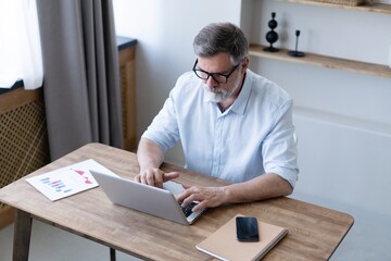 Handsome mature man in casual suit sitting at the table in home office and working at laptop.