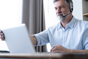 Handsome casual businessman talking online via headset. Mature man having a video call with a laptop at home office - obrazy, fototapety, plakaty