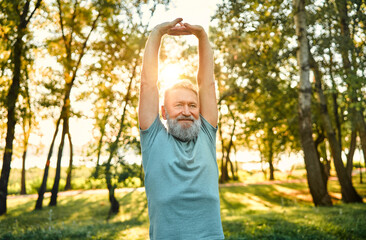 Active and healthy retirement. Portrait of elderly man stretching arms and back before morning exercises on fresh air. Caucasian person warming up while preparing for cardio workout at summer park. - obrazy, fototapety, plakaty