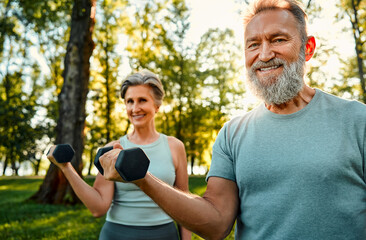 Sport equipment for training. Smiling caucasian man and woman on retirement lifting black dumbbells together outdoors. Fitness married couple working out with weights during summer activity on nature.