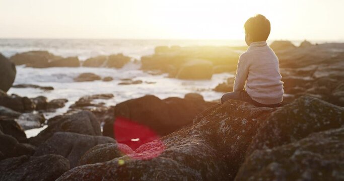 Thinking, young kid and sitting on beach rock by the sea watching a wave at sunset. Child, idea and contemplation in nature with water by the ocean outdoor on holiday feeling relax and calm youth