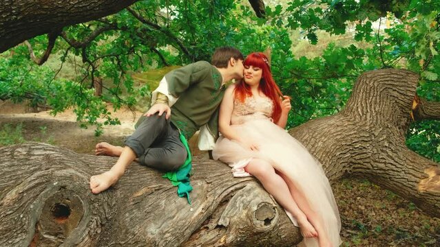 Romantic fantasy couple in love sits on branch old huge oak tree. Elf prince man and fairy woman pixie hugging. Girl happy princess long pink white dress wedding day costume wings. green forest leaves