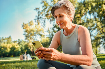 Sport and gadgets. Happy grey haired woman wearing sport clothes sitting at sunny park with modern...