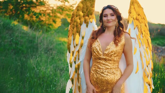 Fantasy happy woman angel beauty golden white wings creative design costume walking through field green grass top hills. Gold color dress. Fairy girl red hair beauty face. sexy lady. diadem on head 4k