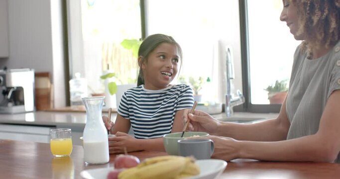 Happy biracial mother and daughter eating breakfast cereal talking in sunny kitchen, slow motion